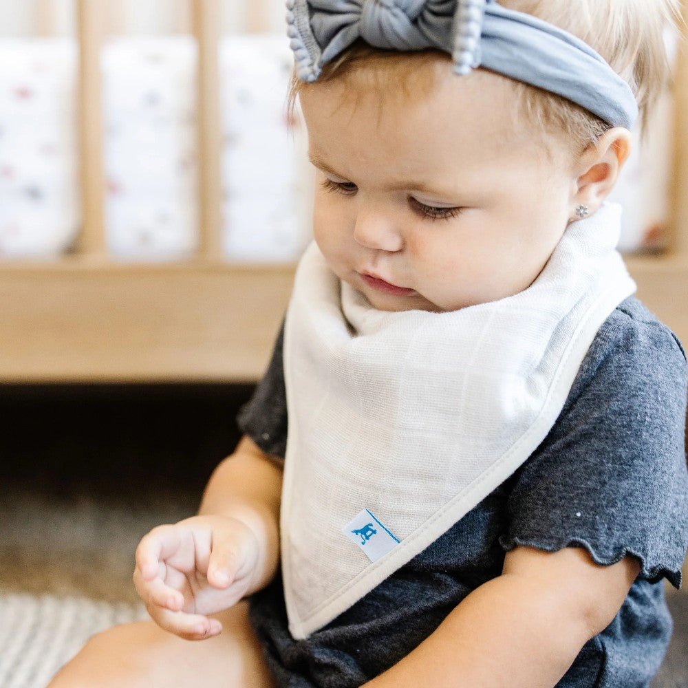 Baby wearing a cream bandana bib and gray headband sitting next tp a wooden baby cot