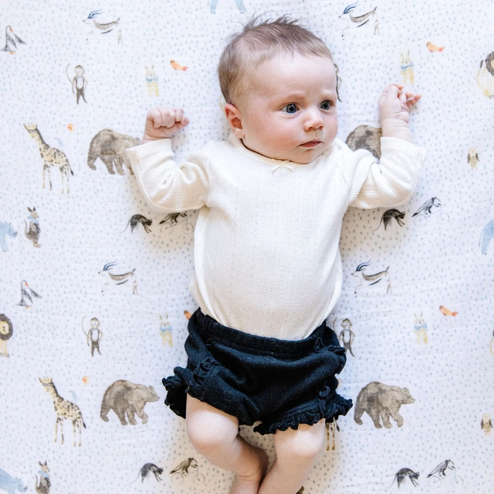 Baby in a white long-sleeve shirt and dark shorts lying on a white and blue dot with animal patterned blanket