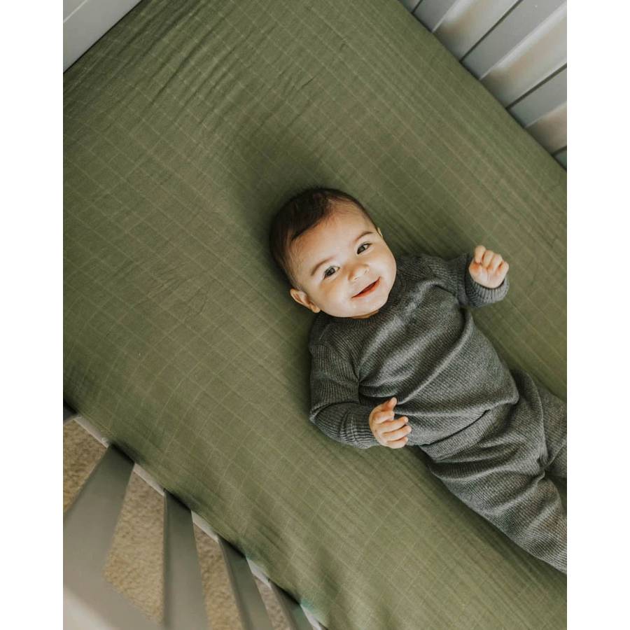Baby wearing a greent rubbed outfit, lying on a cotton muslin cot sheet in green, smiling.