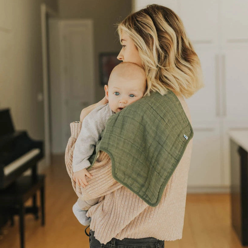 Woman holding a baby with green cotton muslin burp cloth on her shoulder in a room with a piano.