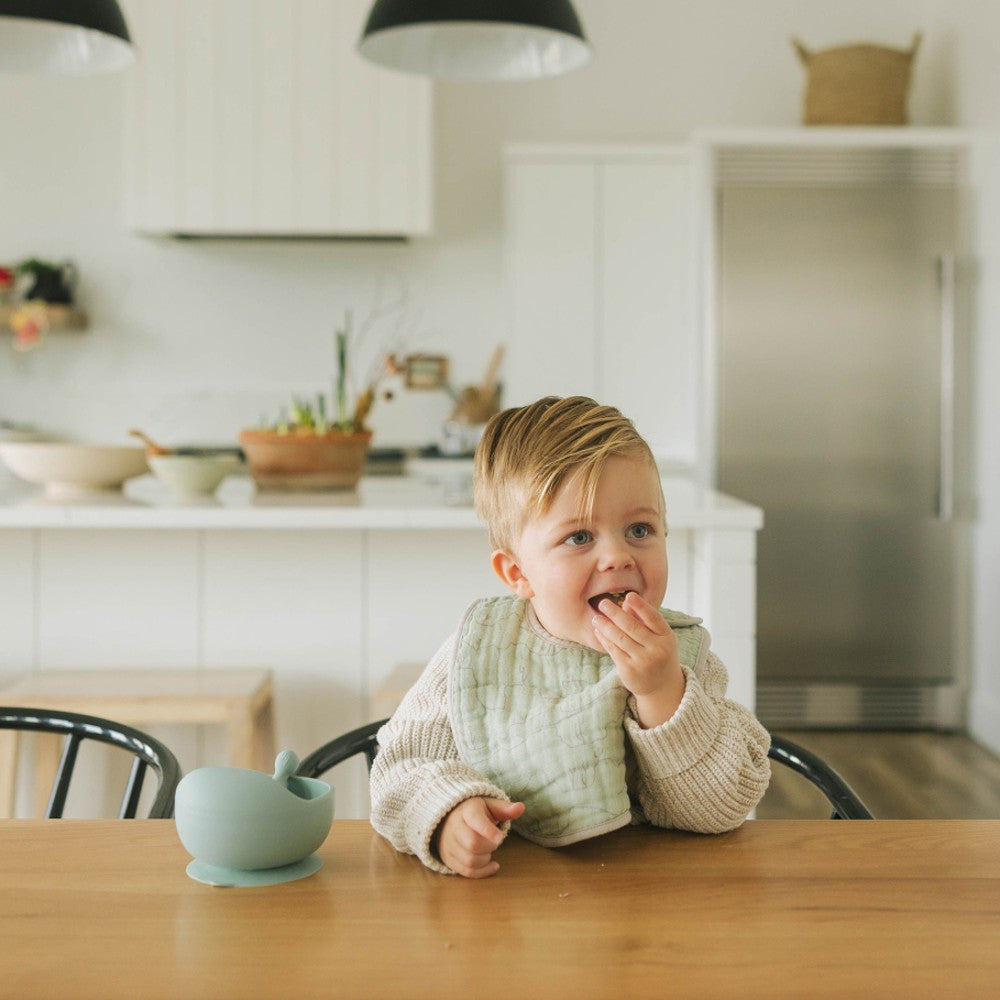 Child sitting at a kitchen table eating, wearing a sage green cotton muslin bib with a blurred kitchen background