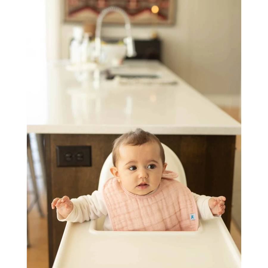 Baby in a high chair with a light pink cotton muslin bib, sitting in a kitchen.