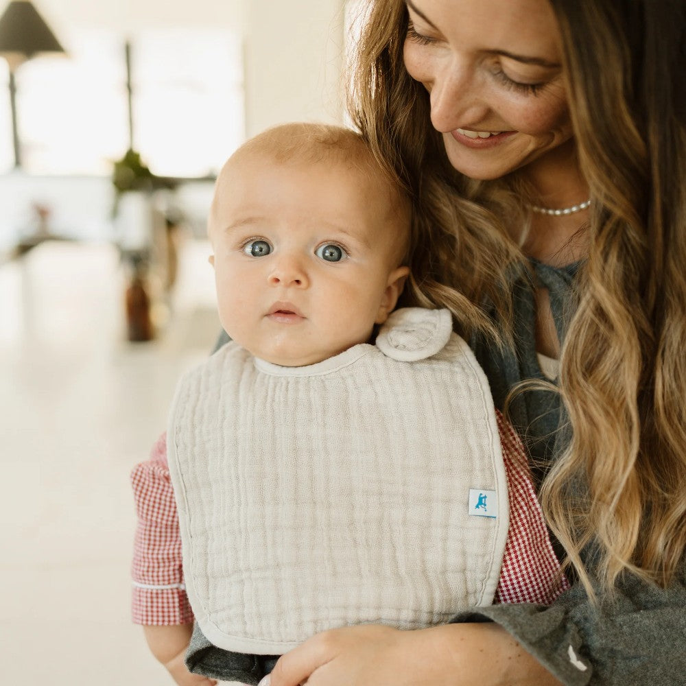 Woman holding a baby wearing a beige/cream classic cotton muslin bib indoors