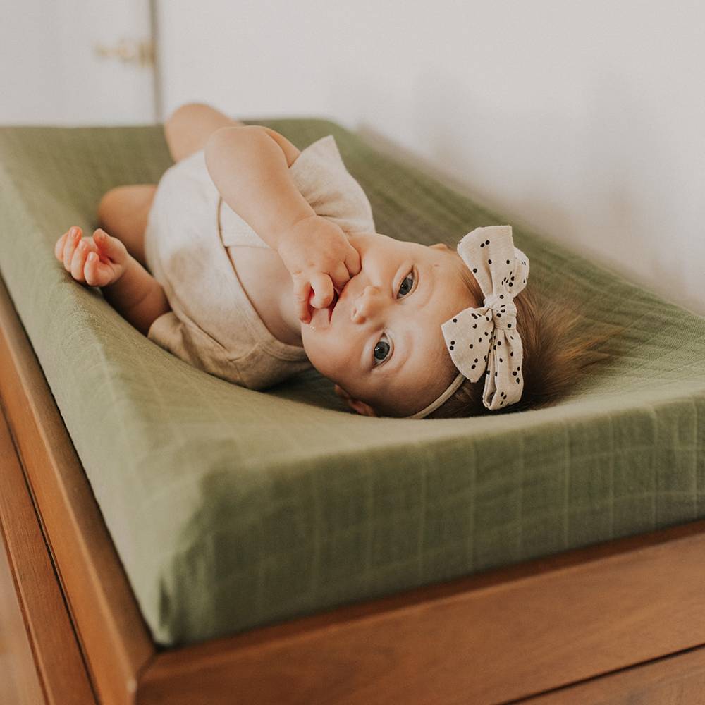 Baby lying on a green changing pad cover on a wooden changing table with a white bow headband.