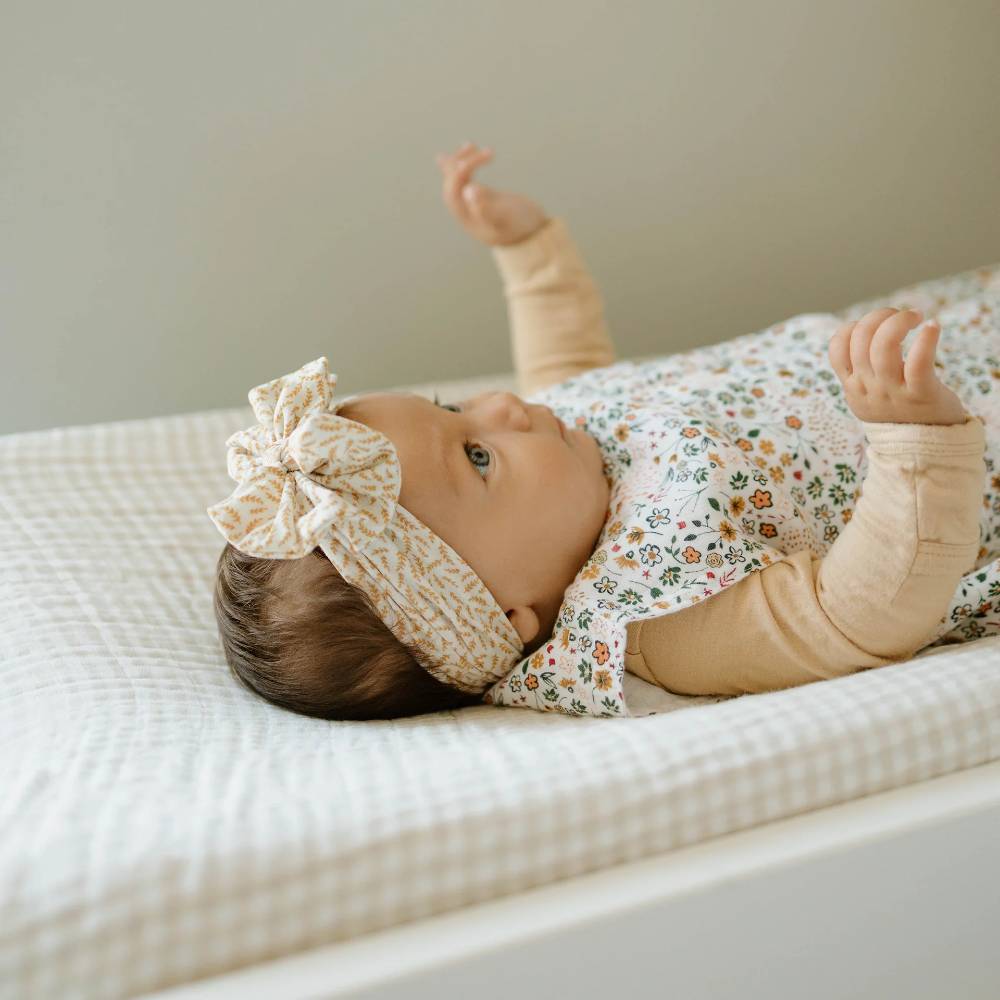 Baby lying on a tan gingham changing pad cover on a changing table wearing a floral outfit and headband.