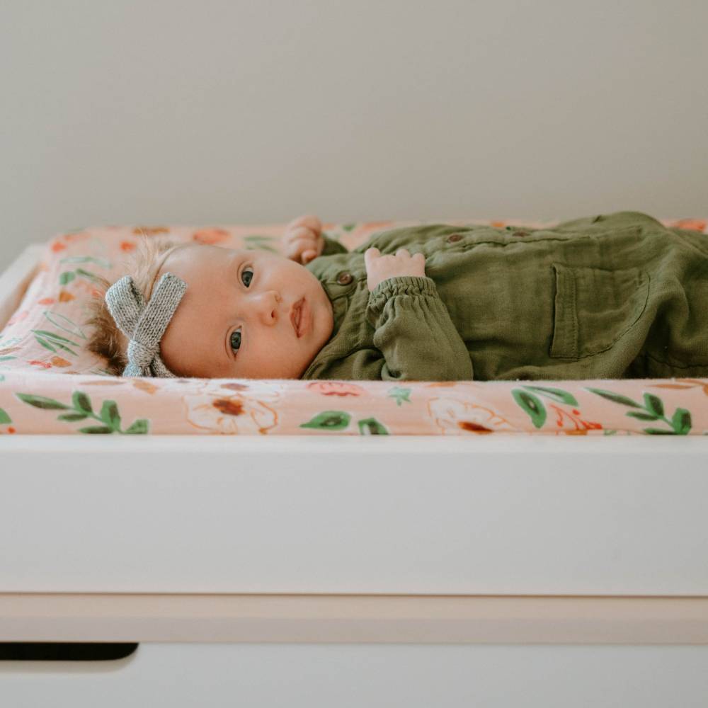 Baby lying on a pink floral changing pad cover wearing a green outfit and gray bow.