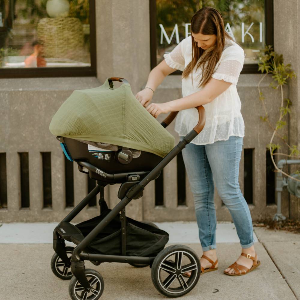 Woman adjusting a green little unicorn cotton muslin car seat canopy on a stroller outdoors.