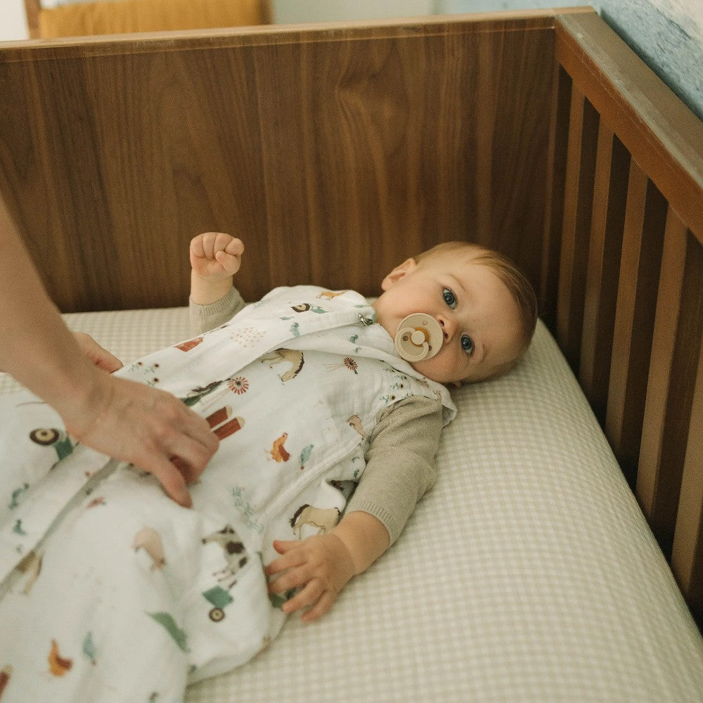 Baby lying in a wooden cot with a beige dummy, wearing a white with farmyard pattern muslin sleeping bag on a tan gingham sheet 