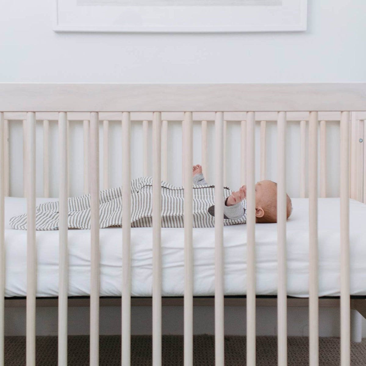 Baby lying in a cream coloured cot with white bedding wearing a grey and white striped muslin sleeping bag 