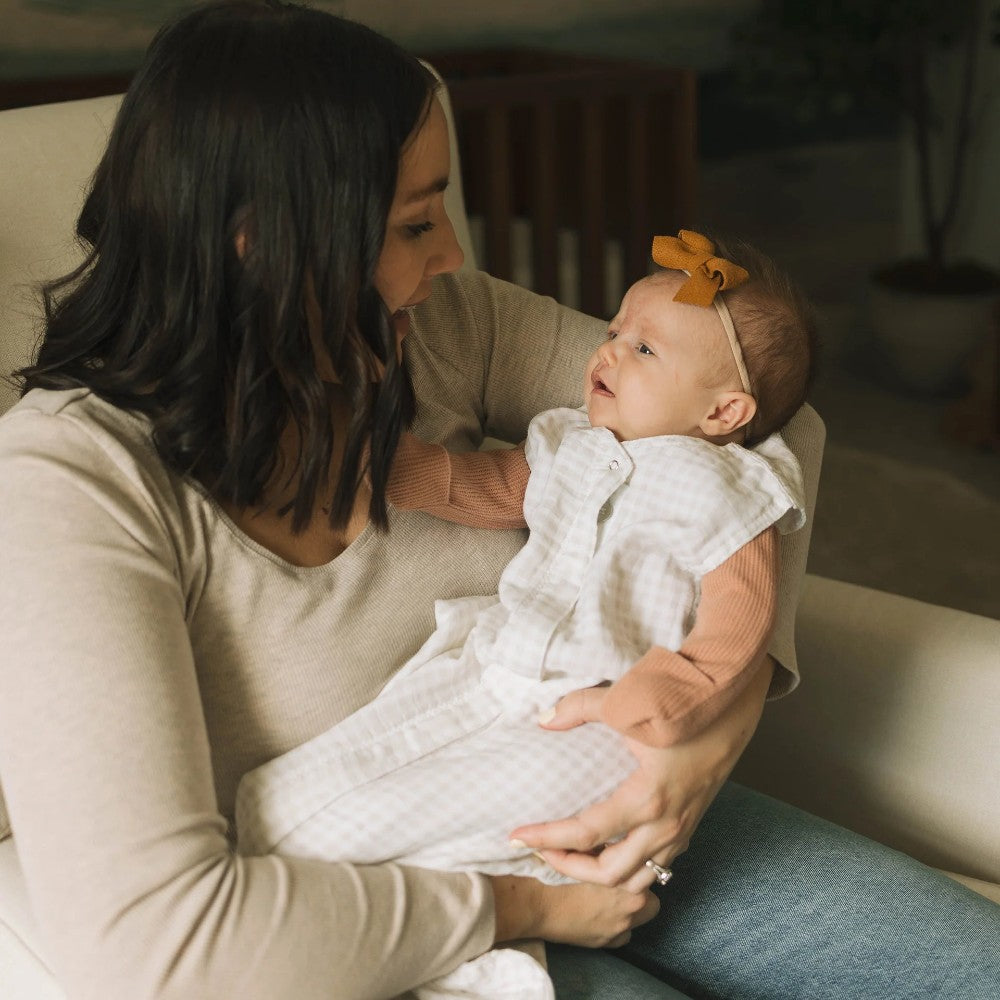 Woman holding a baby in a tan gingham cotton muslin sleeping bag with a bow in a blurred indoor setting