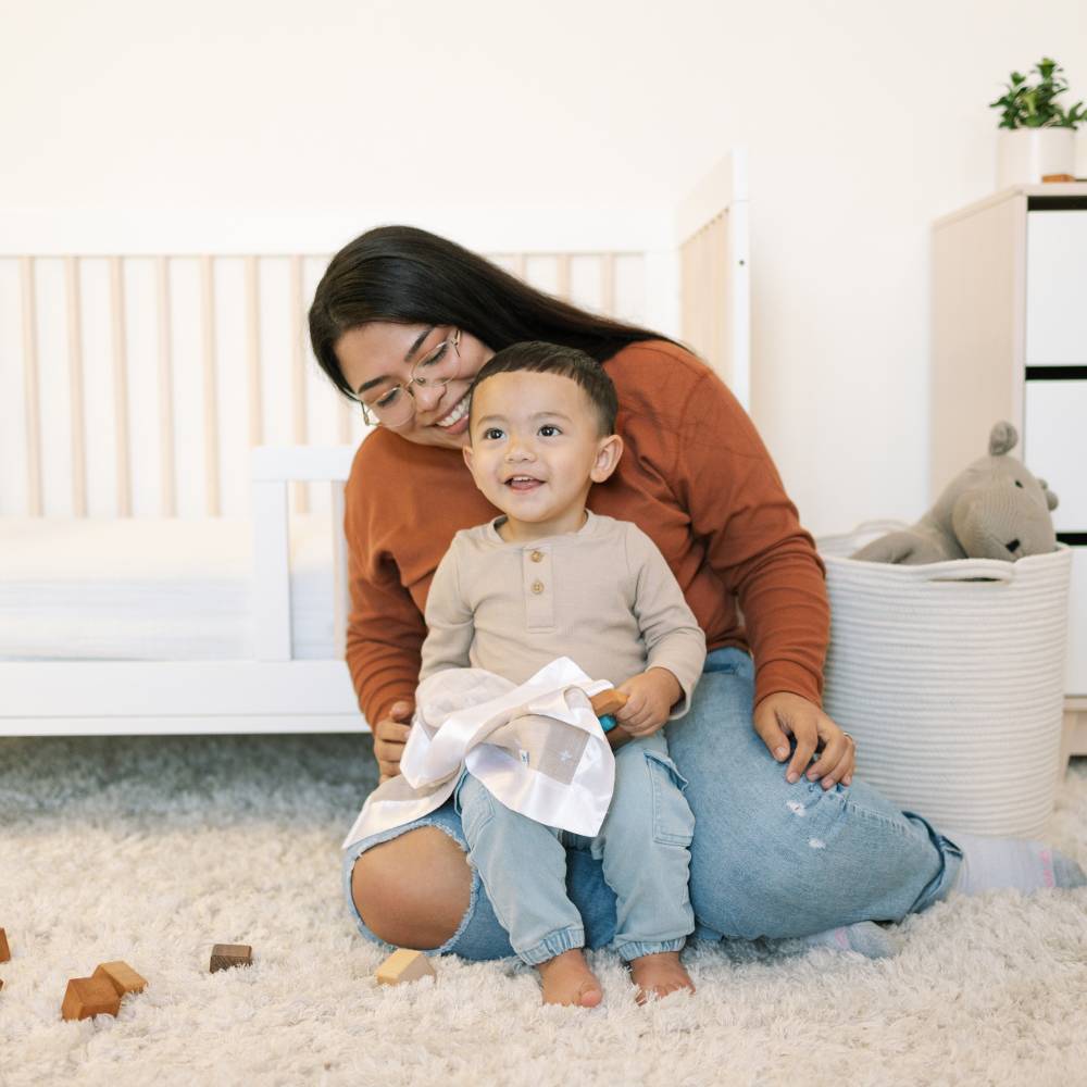 Woman and child sitting on a carpeted floor in a home setting holding a beige with crosses muslin cotton security blanket with white satin trim 