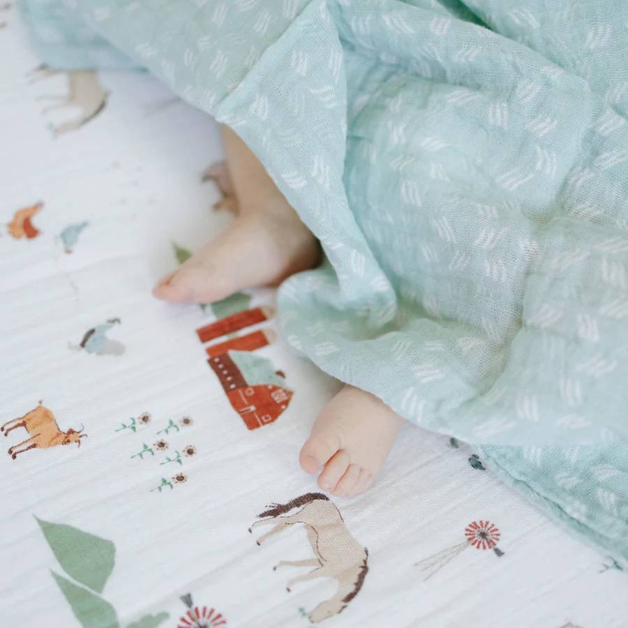 Baby feet peeking out from under a light blue cotton muslin blanket on a patterned surface