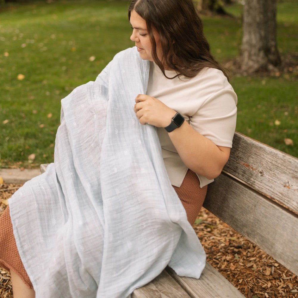 Woman sitting on a bench with a light blue muslin swaddle blanket wrapped around her in a park setting.