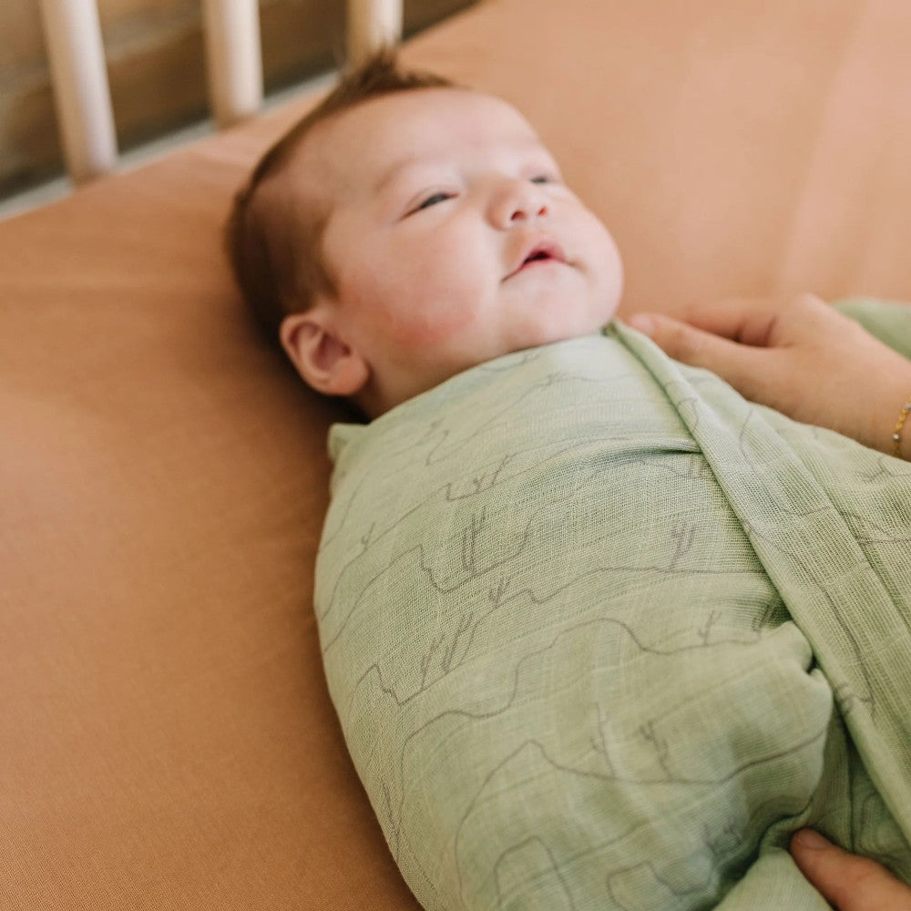 Baby wrapped in a green cotton muslin swaddle blanket lying on a rust coloured sheet