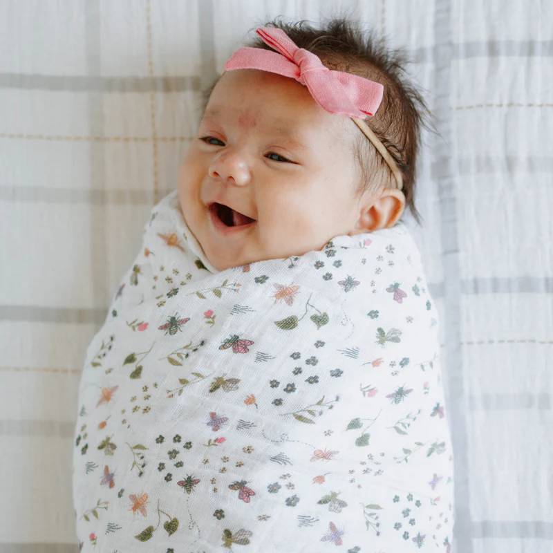 Baby wrapped in a floral garden themed muslin swaddle blanket with a pink headband, smiling against a neutral background