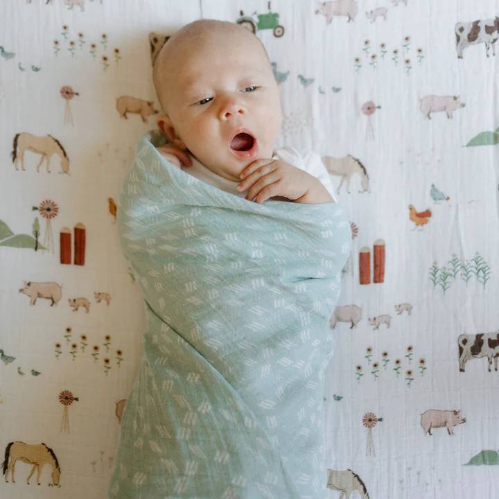 Baby wrapped in a light green swaddle blanket with a farm animal pattern in the background