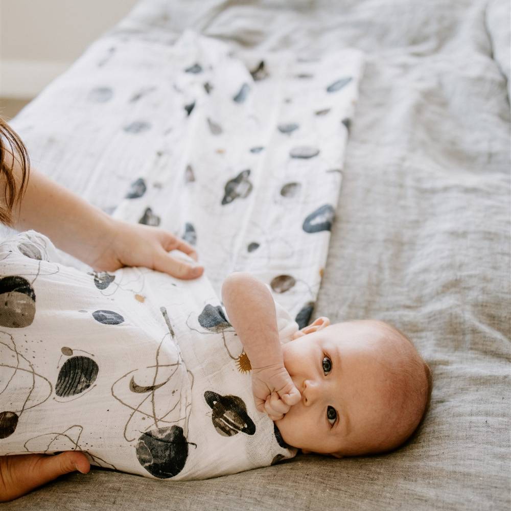 A baby lying on a bed and wrapped in a planet themed white cotton muslin swaddle wrap
