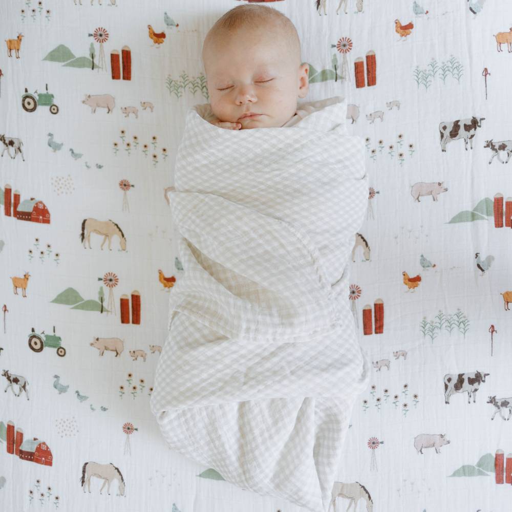 Baby wrapped in a tan gingham blanket on a cot sheet with farm animal patterns