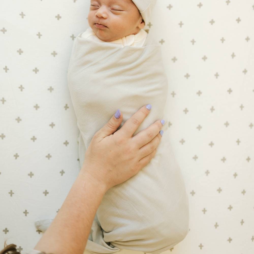 Newborn baby swaddled in a pebble grey blanket with a hand holding it, on a patterned background