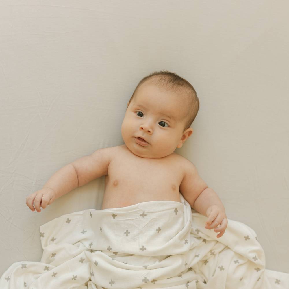 Baby wrapped in a white blanket with cross patterns on a beige stretch sheet background