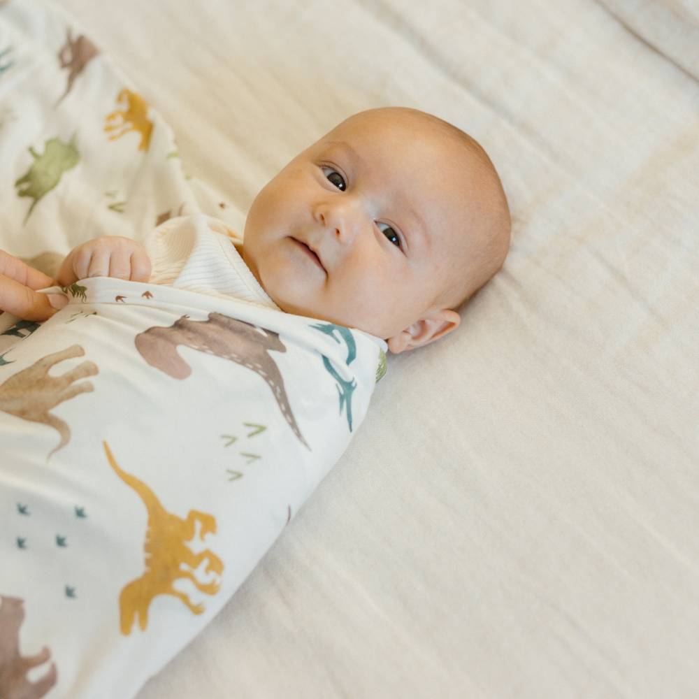 Baby wrapped in a knit stretch swaddle blanket with dinosaur pattern on a soft surface