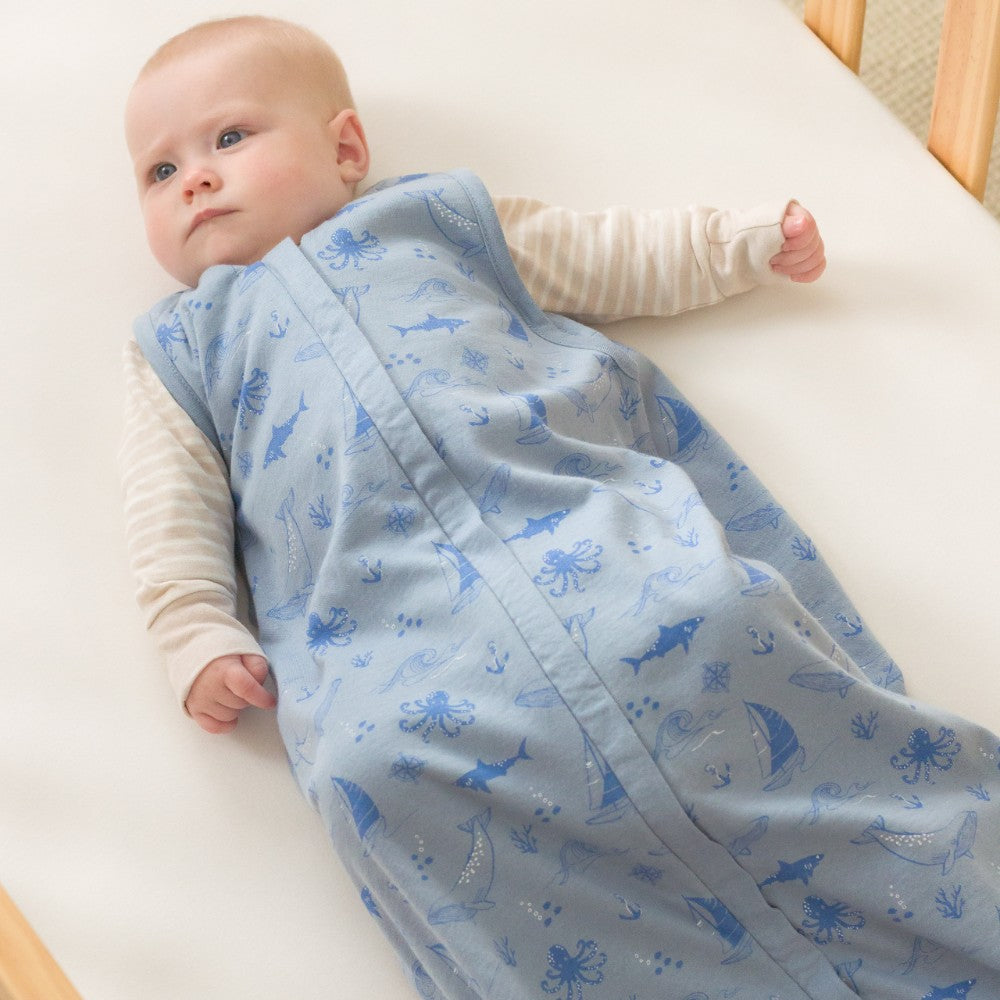 Baby wrapped in a blue sleep sack with sea creature patterns on a white surface.