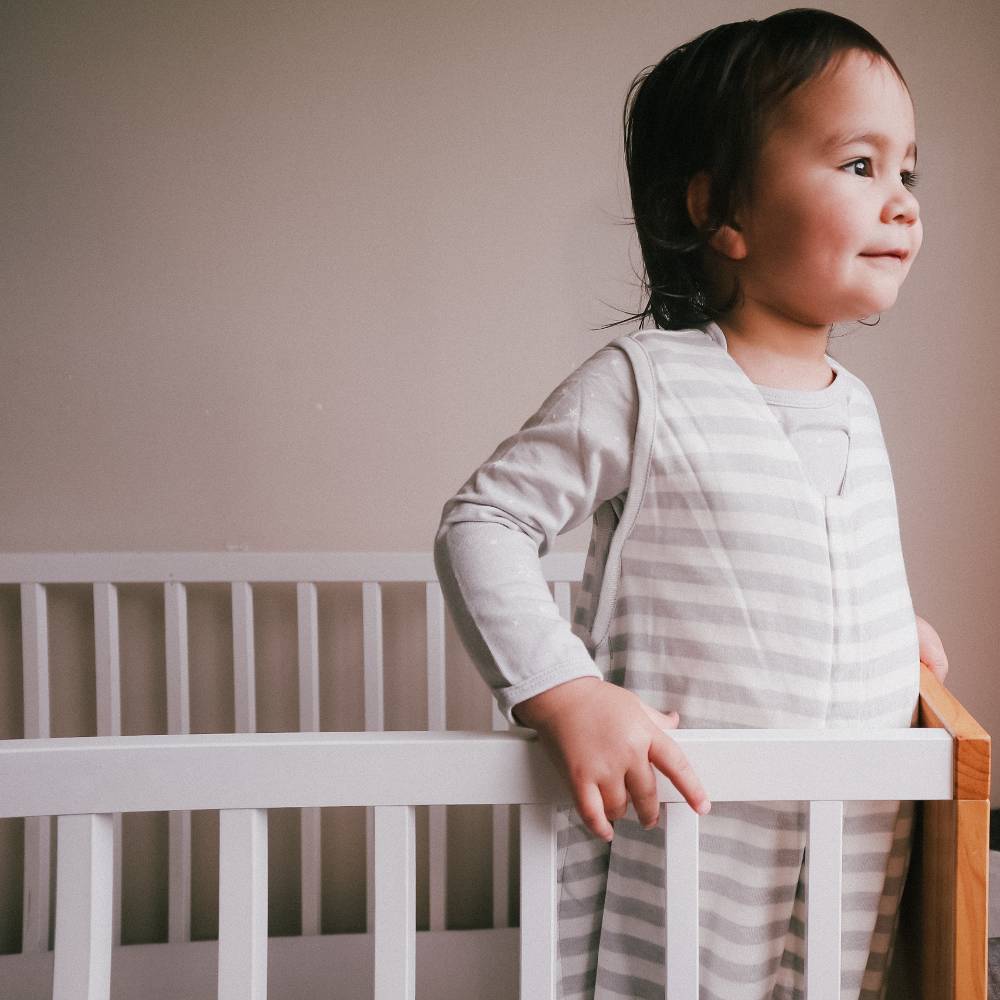 Child standing in a crib wearing a striped outfit against a plain wall.