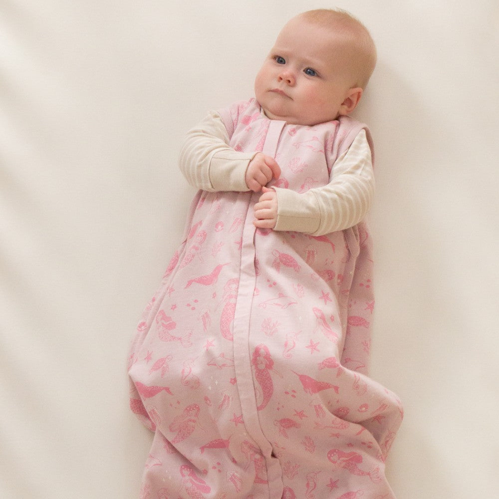 Baby lying on a white surface wearing a pink mermaid print woolbabe sleeping bag