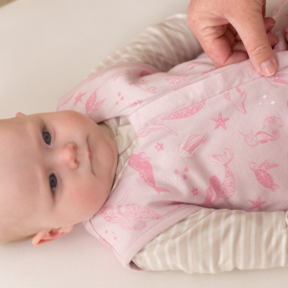 Baby wearing a pink outfit with mermaid patterns, lying on a white surface.