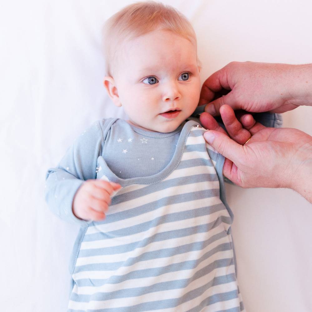 Baby wearing a blue and white striped outfit with a person adjusting it on a light background