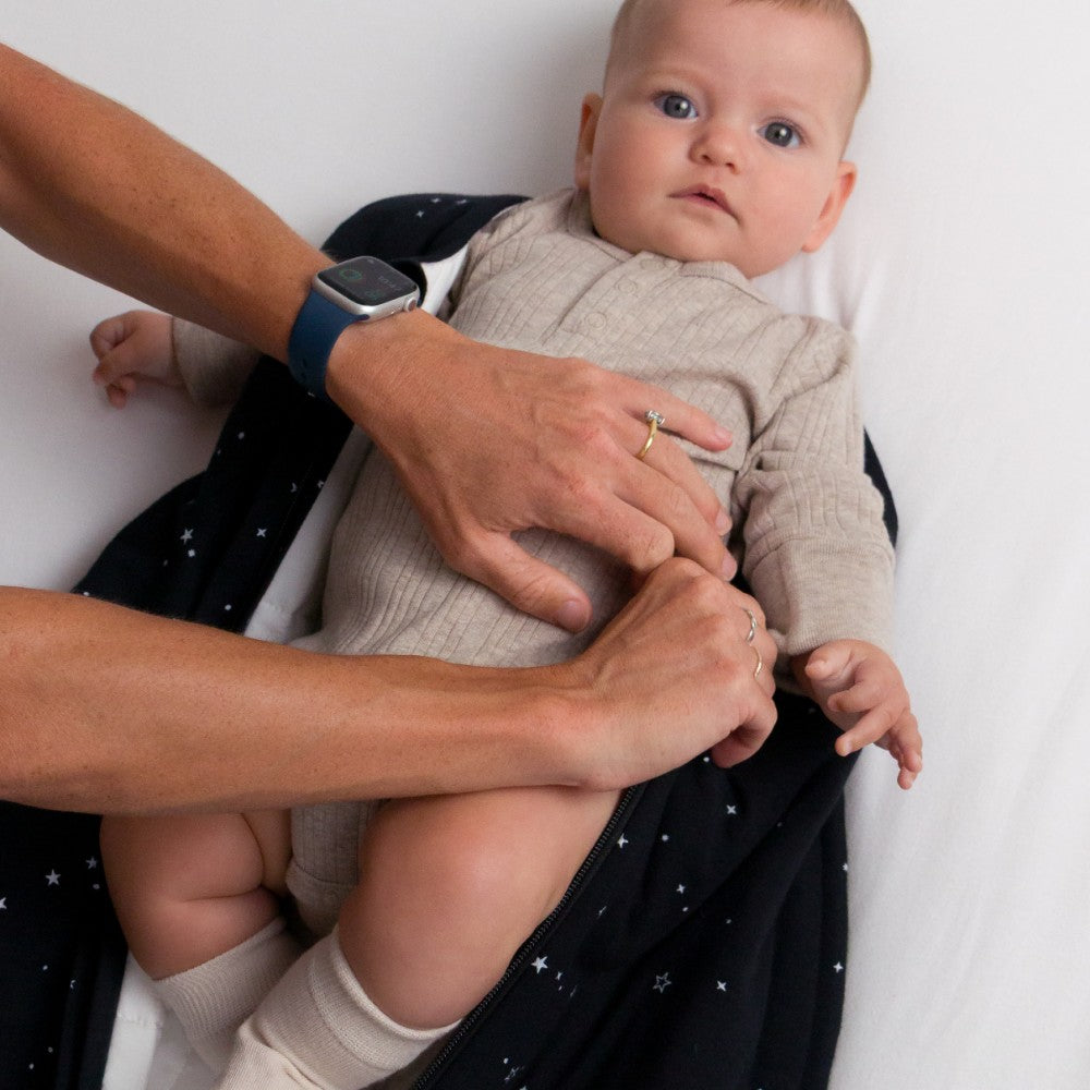 Baby wewaring a beige Dune marl bodysuit in a woolbabe mini sleeping bag with adults hands dressing the baby