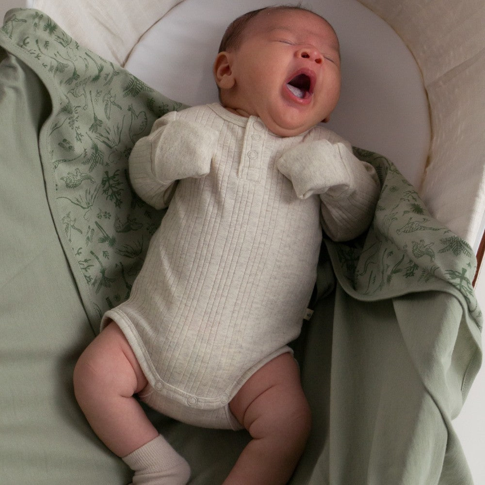 Newborn baby in a beige rib bodysuit yawning in a crib with green bedding.