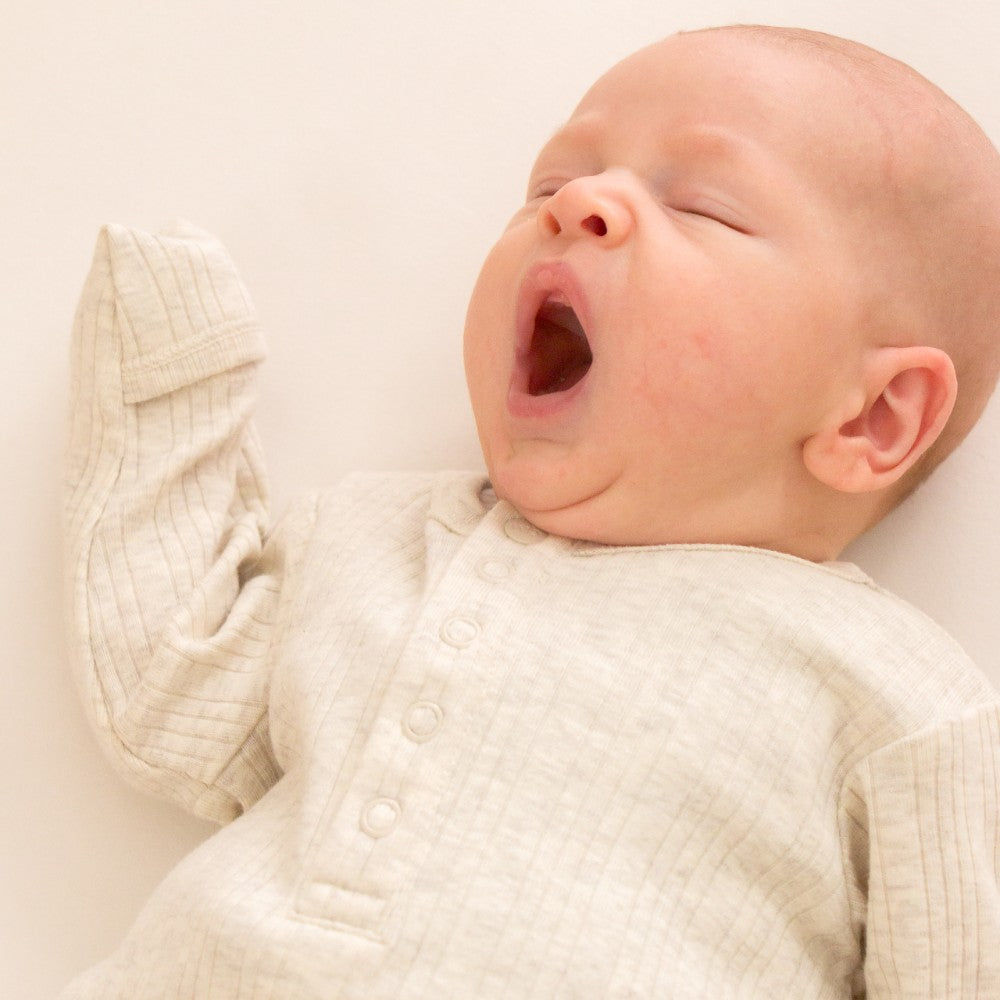 Baby in a woolbabe gown with hands in foldover mittens in a textured outfit yawning on a plain background