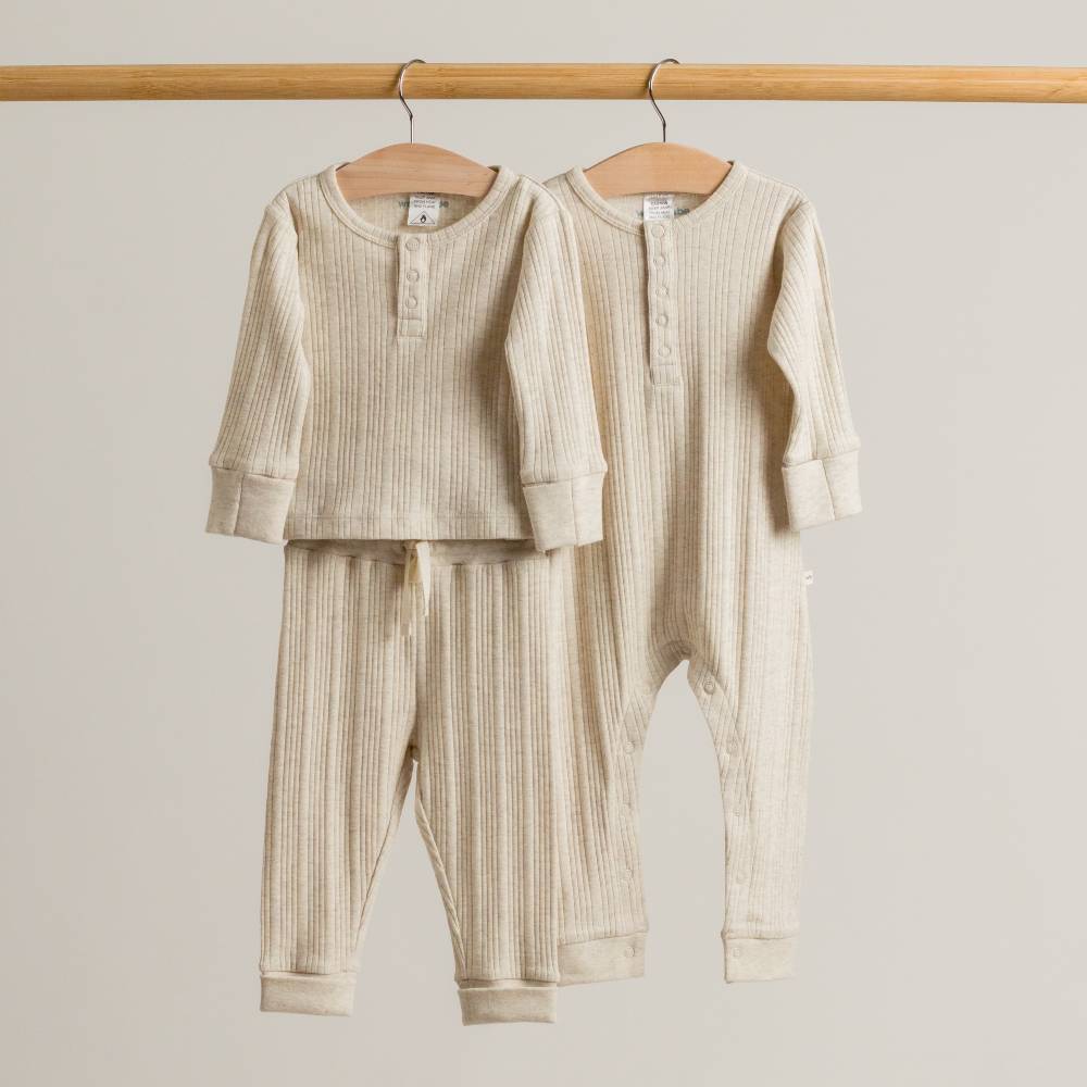 Two cream marl woolbabe baby outfits hanging on a wooden rod against a plain background