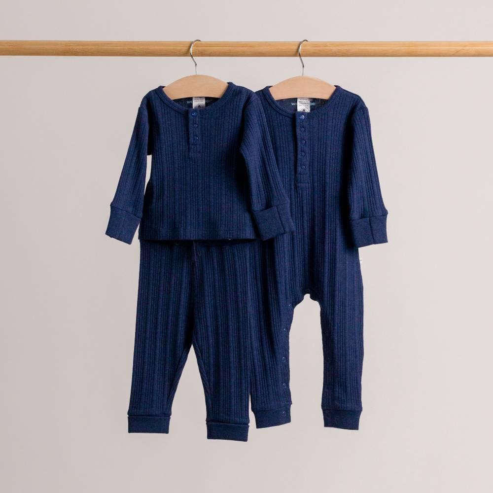 Two navy ribbed woolbabe daydream lue children's outfits hanging on a wooden rod against a light gray background.