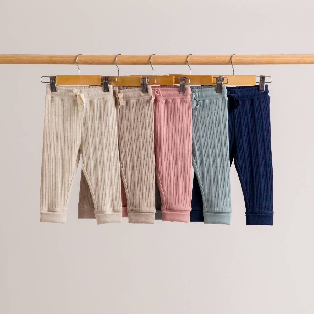 Five pairs of woolbabe rib drawstring daydream pants in different colors hanging on a rack against a plain background