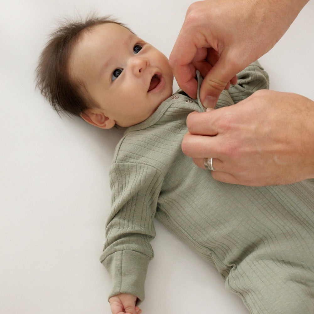 Baby wearing a green woolbabe onesie with adult hands adjusting it on a white background