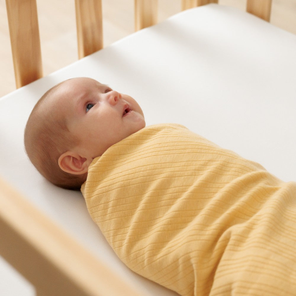 Newborn baby swaddled in a buttercup yellow woolbabe swaddlle blanket lying on a white surface.