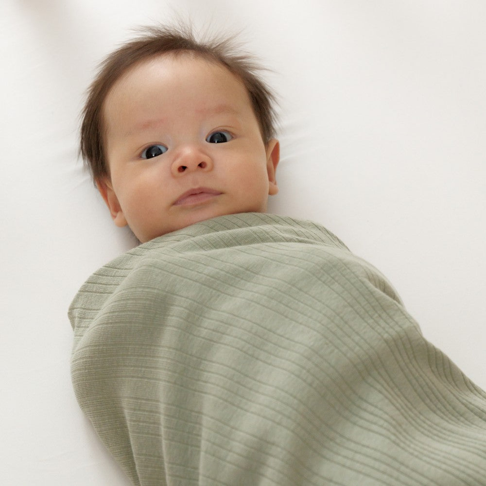 Newborn baby wrapped in a green rib merino swaddle on a white background