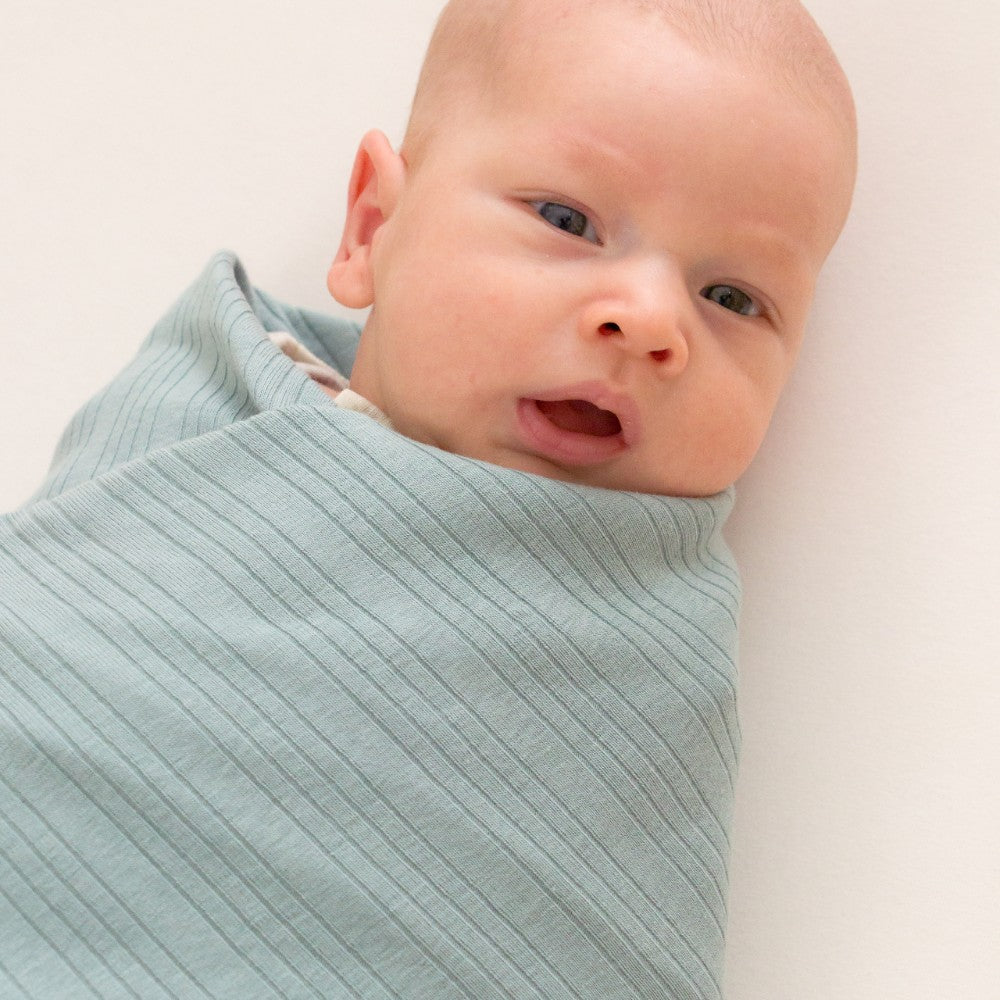 Baby wrapped in a light blue woolbabe merino swaddle on a white background