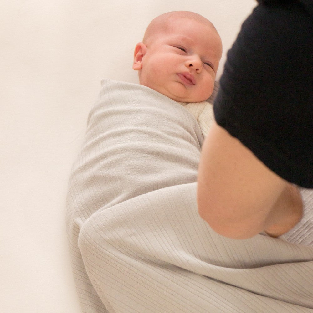 Newborn baby wrapped in a light gray swaddle blanket with a blurred person in the background.