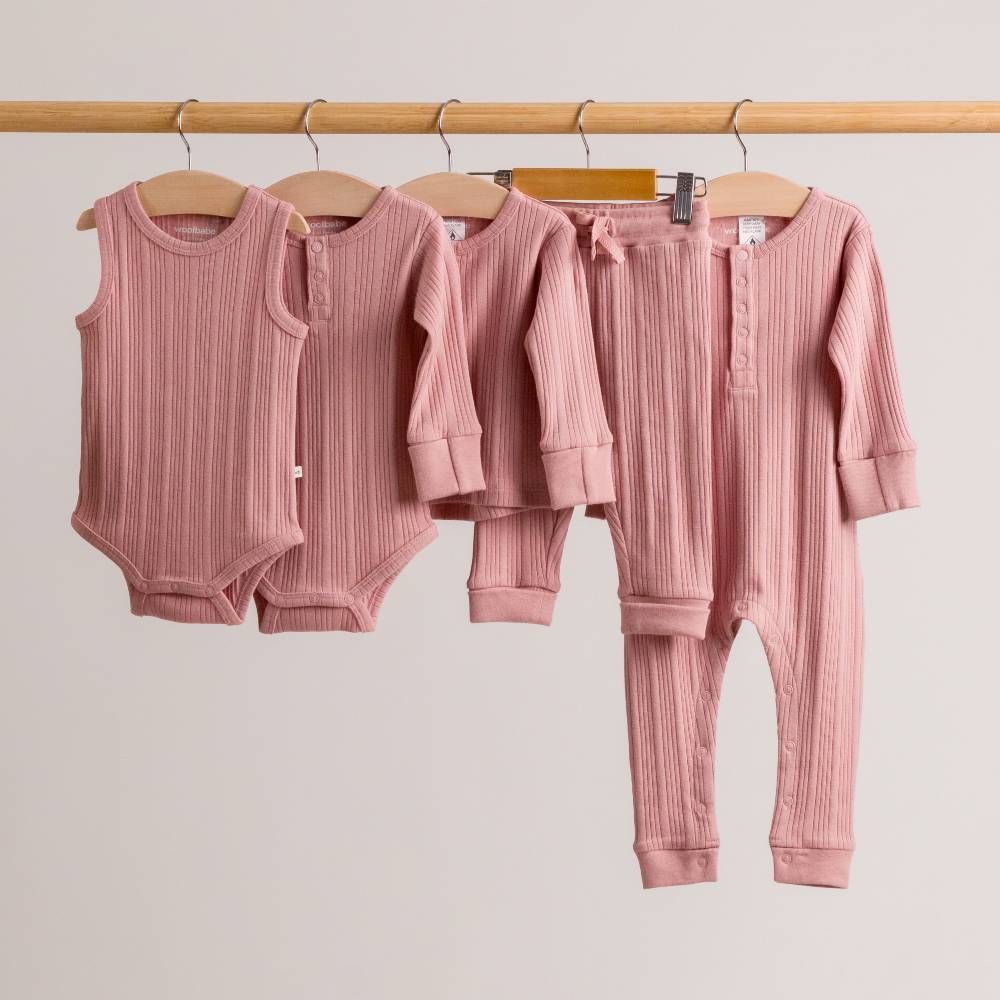 Set of pink woolbabe rib baby clothing including rompers and pants hanging on a rack against a plain background.
