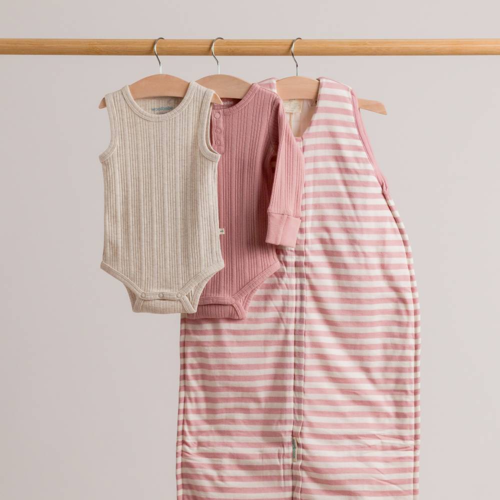 Baby clothing including a sleeveless onesie and striped pants on hangers against a plain background.