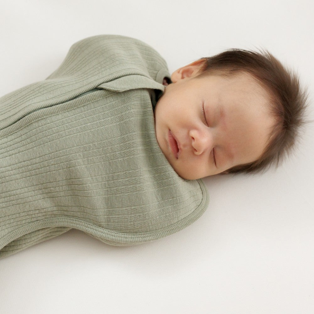 Newborn baby swaddled in a green woolbabe merino zip swaddle on a white background
