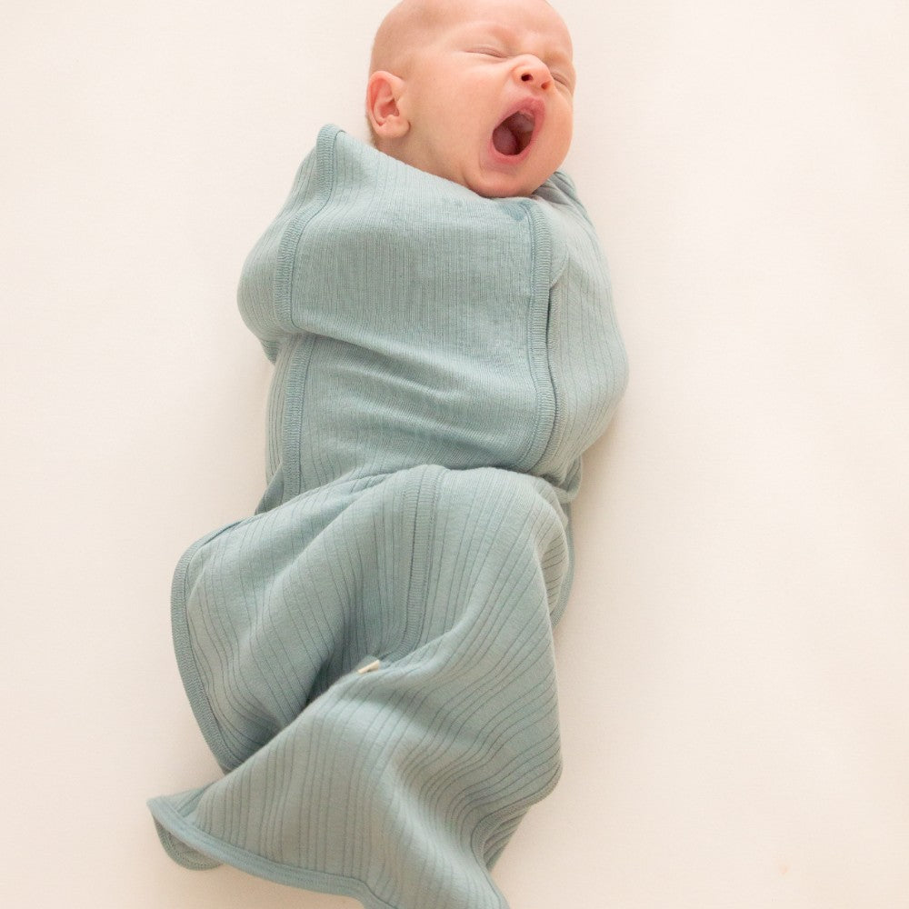 Newborn baby wrapped in a light blue woolbabe merino zip swaddle on a white background