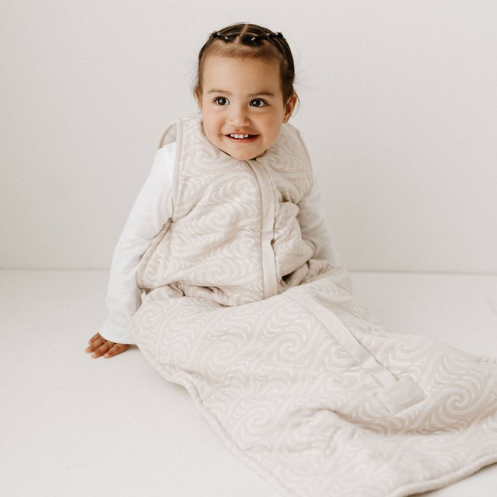 Child wearing a white quiltedsleeping bag with maori print on a plain background