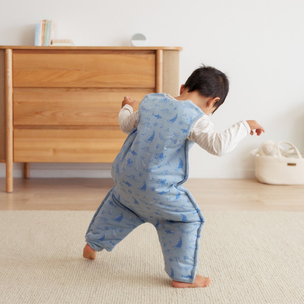 Child in a blue patterned outfit standing on a beige carpet with a wooden dresser in the background.