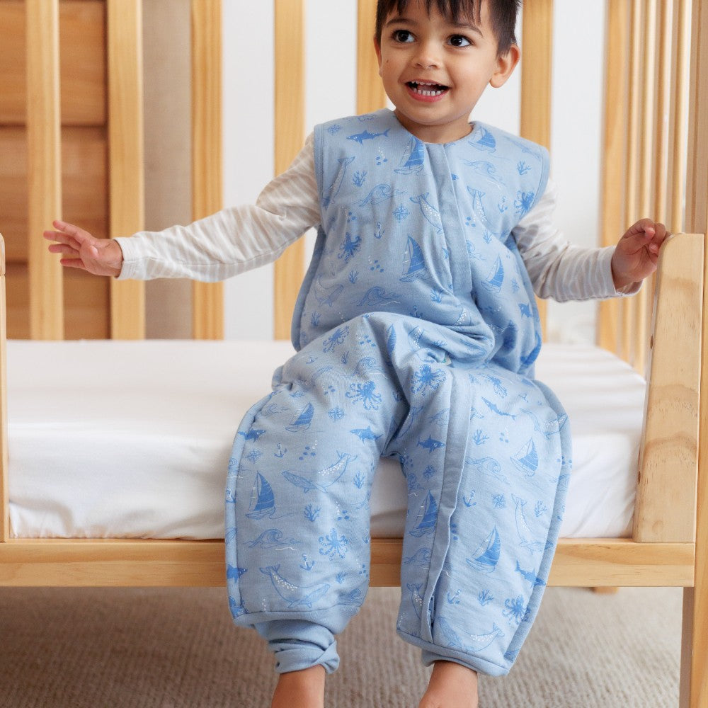 Child wearing a blue patterned winter sleep onesie sitting on a wooden crib.