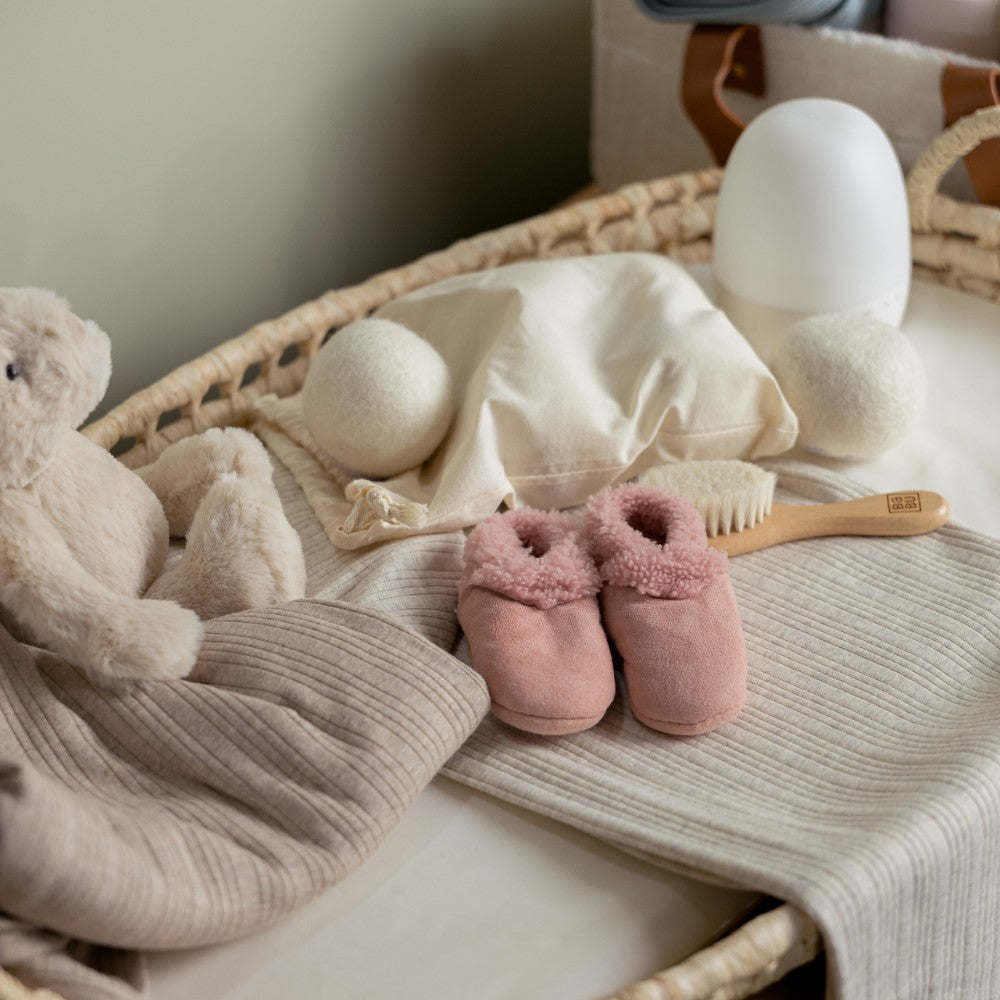 Baby items including pink slippers, a teddy bear, and a basket on a light surface.