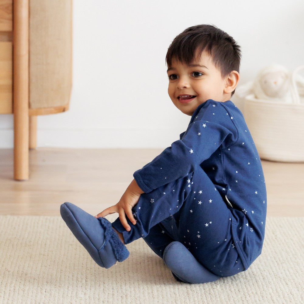 Child wearing a blue woolbabe pyjamas and navy blue lambskin slippers with star patterns sitting on a beige rug.